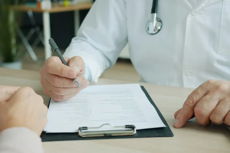 Doctor writing on a form on a clipboard, with a patient's hands resting on the desk opposite