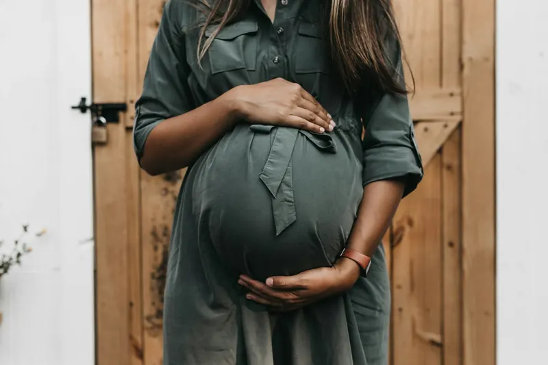 Pregnant woman in a green shirt dress holding her belly in front of a wooden door